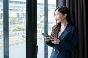 Young businesswoman using smartphone working in hotel room during her business travel. Businesswoman working from hotel room on business trip standing by the window in sunny day.