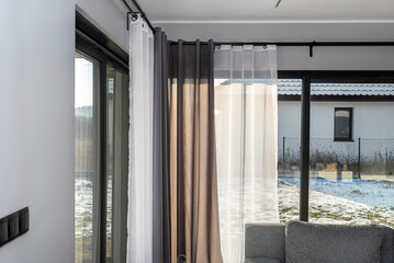 A modern and simple living room in the house with large terrace windows with curtains and a gray...