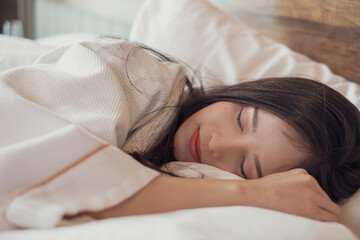 Young Asian woman with lying in bed under white cover in the morning. Young pretty lady sleeping in bed. Young woman on bed in modern apartment. Woman stretching in bed after wake up.
