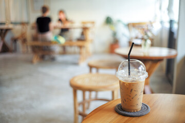 Closeup plastic glass of ice coffee on the wooden table with blurred customers in background. Co working space concept