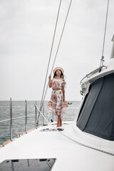 Sublime young lady in a beach dress and panama hat is standing and posing on her private yacht, portrait