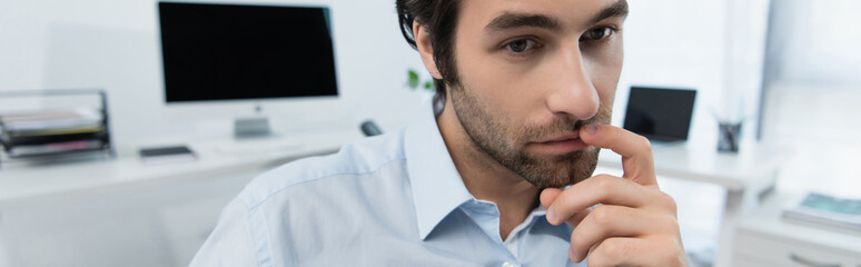 pensive businessman touching lips while thinking near computers on blurred background, banner.