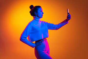 Portrait of young beautiful girl using phone isolated on orange background in neon light, filter. Concept of emotions, facial expression, youth, aspiration, sales - obrazy, fototapety, plakaty