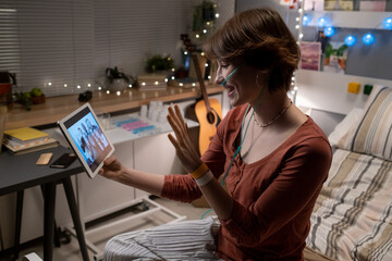 Happy teenage girl looking at her friends on tablet screen and waving hand to them during...