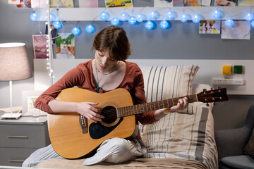 Teenage girl playing guitar while sitting on bed in hospital ward and taking online course of music education