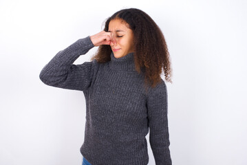 teenager girl wearing grey sweater standing against wite background smelling something stinky and...