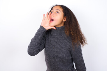 teenager girl wearing grey sweater standing against wite background shouting and screaming loud to side with hand on mouth. Communication concept.