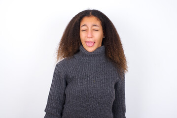 teenager girl wearing grey sweater standing against wite background sticking tongue out happy with funny expression. Emotion concept.