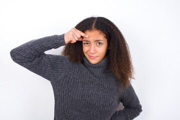 teenager girl wearing grey sweater standing against wite background pointing unhappy at pimple on forehead, blackhead  infection. Skincare concept.