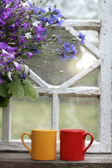 red and yellow mugs next to bouquet of wild flowers on the windowsill against the backdrop of retro window. morning coffee in the village
