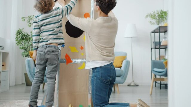 Slow motion of family mom and son making cardboard spaceship decorating with stickers at home. Leisure activity and happy childhood concept.