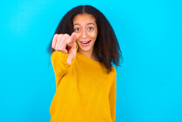 Excited positive beautiful teenager girl wearing yellow sweater standing against blue background points index finger directly at you, sees something very funny. Wow, amazing