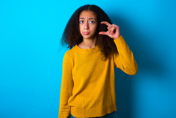 beautiful teenager girl wearing yellow sweater standing against blue background purses lip and gestures with hand, shows something very little.