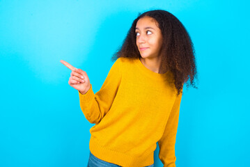 beautiful teenager girl wearing yellow sweater standing against blue background  points at copy space and advertises something, advices best price.