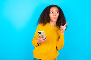 Stunned beautiful teenager girl wearing yellow sweater standing against blue background points...