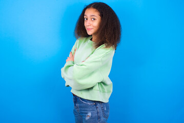 Fototapeta na wymiar Portrait of beautiful teenager girl wearing green sweater standing against blue background standing with folded arms and smiling