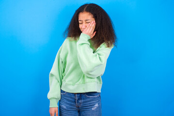 beautiful teenager girl wearing green sweater standing against blue background with toothache
