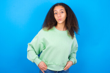 Portrait of lovely funny beautiful teenager girl wearing green sweater standing against blue background sending air kiss