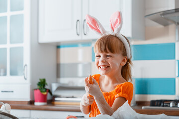 Emotional portrait of cheerful little girl dressed as bunny with carrot for Easter while cooking food in kitchen at home. Girl kid child having fun laughs plays with flour and carrot.