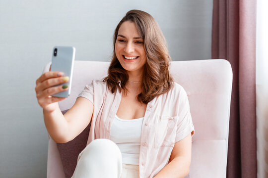 Beautiful tender young smiling caucasian 30s woman taking picture selfie using cell phone. Long haired brunette is photographing yourself on smartphone home interior.