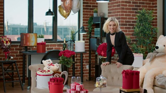 Surprised beautiful woman finds room full of valentine day gifts and roses hugs teddy bear feeling happy. Seductive blonde recieving romantic luxury gifts celebrating love in festive decorated living.