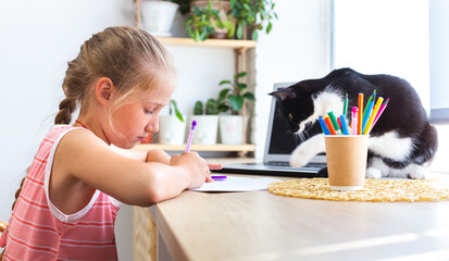 Child writing or drawing in notebook. A black and white cat disturb girl from doing her homework. Caucasian cute girl doing homework at home, next to laptop. Cozy workplace by window with houseplants.
