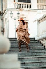 Outdoor fashion portrait of young elegant fashionable brunette woman, model in stylish hat, choker and light raincoat posing at sunset in European city.