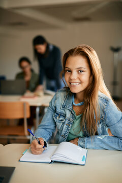 Happy High School Student Writing In Notebook During A Class And Looking At Camera.