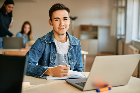 Smiling teenager taking notes while using laptop during a class at high school.