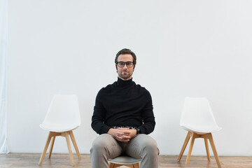 young and stylish man in black turtleneck and eyeglasses sitting on chair and looking at camera.