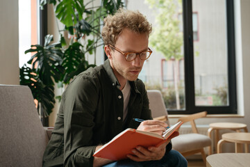 Pensive student studying, learning language sitting at workplace.  Education concept. Portrait of young handsome man writer wearing stylish eyeglasses taking notes working from home    - obrazy, fototapety, plakaty