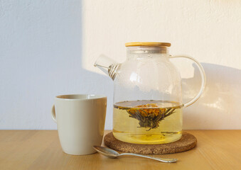 Glass teapot with fresh green tea, beige ceramic cup and tea spoon on a wooden table