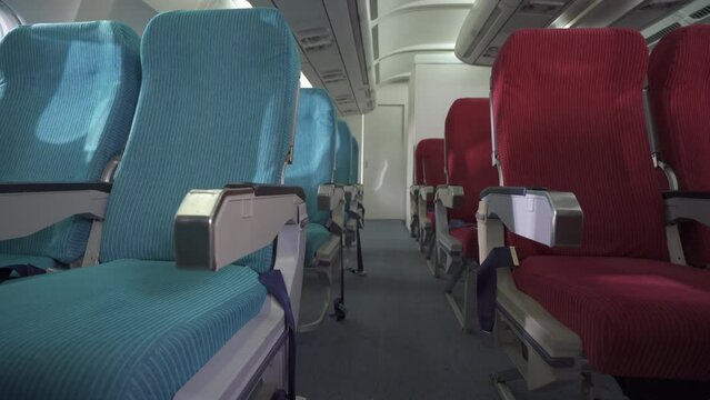 An inside of the airplane on the aisle between rows of an empty blue and red airplane seats with no passenger both sides during daytime. Airline transportation and tourism concept.
