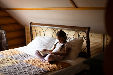 Young serious female kid holding book in hands reading attentively sitting on bed with crossed legs wearing home clothes in wooden house. Free time leisure activities. Self-development.