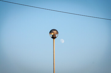 Light pole at sunset in front of a blue sky with the moon, the pole is hit by the last rays of...