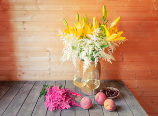 Bouquet of yellow lilies and white astilbe in clay vase, glasses of white wine, pink astilbe and fresh fruits near.
