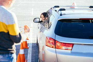 Positive experience in driving school. Cheerful confident young woman is glad for improvement of her driving skills with orange road cone. Male instructor stands near the car holding clipboard.