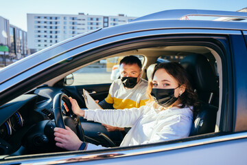 Brunette middle-eastern woman in face mask learning parking at driving school while COVID-19 pandemic, holding hands on automobile steering wheel, sitting by male driving instructor, side view