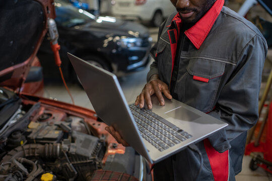Part of repairman in workwear holding laptop while standing by car with open hood and consulting clients online in garage