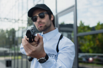 blurred security man in cap and sunglasses holding gun outdoors.