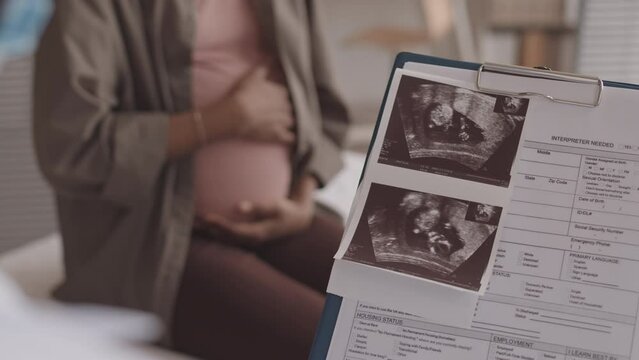 Medium closeup with slowmo of unrecognizable gynecologist looking at baby ultrasound image and patient medical card while talking to pregnant woman sitting on examination couch