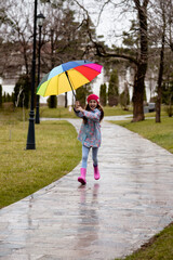 A cute little girl in a blue cape, pink boots and a pink hat runs through puddles and has a fun.The girl has a rainbow umbrella in her hands. Happy childhood. Early spring. Emotions.