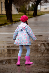 A cute little girl in a blue cape, pink boots and a pink hat runs through puddles and has a fun. Happy childhood. Early spring. Emotions.