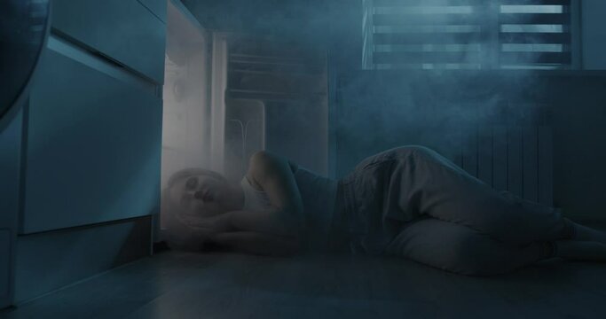 A young woman is sleeping by the open refrigerator. The girl escapes from the heat without air conditioning.