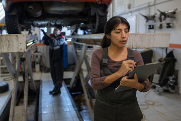Young pretty female worker of car service making notes in document on clipboard against her male colleague repairing vehicle