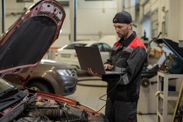 Serious young repairman with laptop making technical diagnostics of diesel engine of car while...
