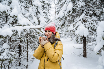 Fototapeta na wymiar A girl holds a mug of hot tea in her hands. A woman on a walk in the winter forest drinks tea. Red hat on the head. Yellow jacket. Thermo glass in hand.