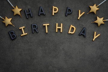 Happy birthday letters in shiny gold and matte black color on a gray background, golden candles in the form of stars