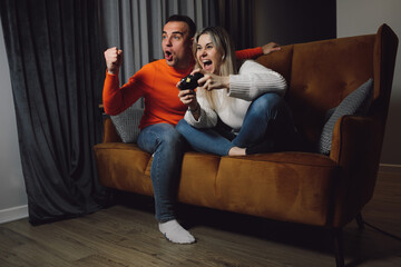 Dark-haired smiling enthusiastic man and blonde woman winner playing video games joystick TV on couch indoor