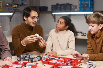 Young man in eyeglasses sitting at the table with his students and teaching them robotics